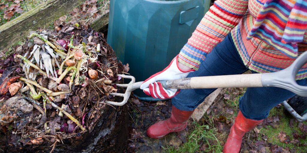 A pile of compost being forked over by a woman in a striped sweater and red wellies. Her face is not visible.
